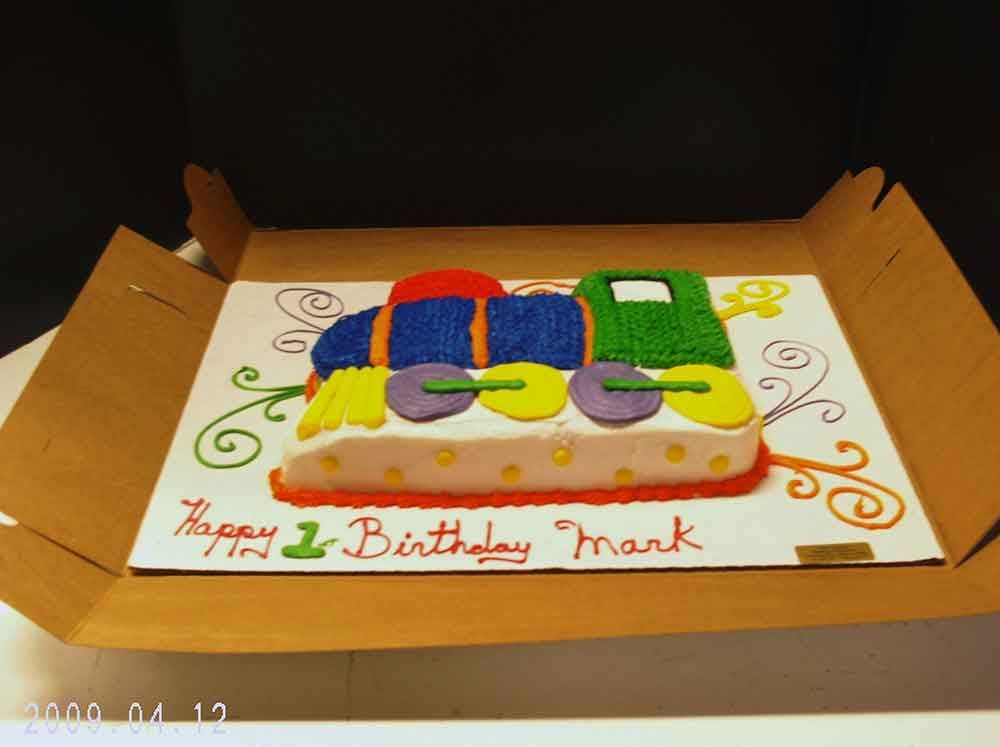 Train-shaped cake with colorful frosting in a cardboard box, reads 