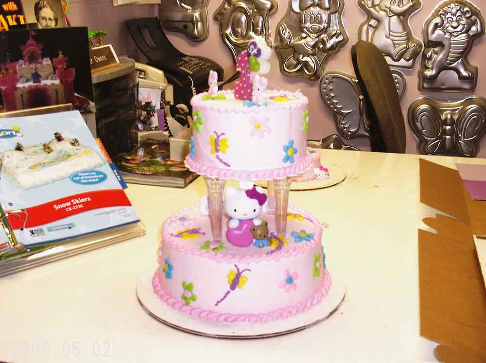 Two-tiered pink cake with Hello Kitty figure, flowers, and butterflies on a white table.