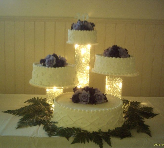 Four-tiered wedding cake display with purple flower accents, lit from within, on a table with greenery.