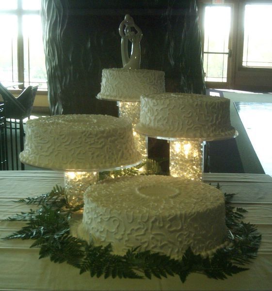 Four-tiered wedding cake with white frosting and a couple topper, displayed on illuminated stands, surrounded by greenery.