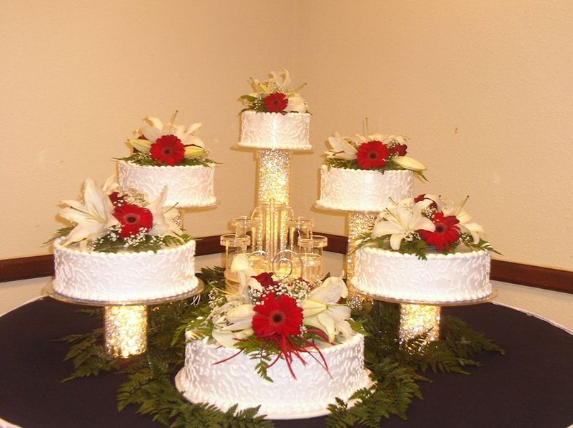 Wedding cake display with five tiers, white frosting, red and white flowers, and lit pedestals.