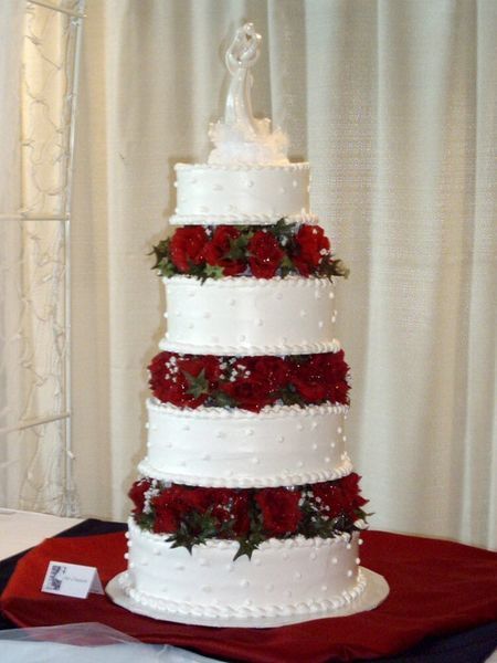 Multi-tiered white wedding cake decorated with red roses and a bride and groom topper.