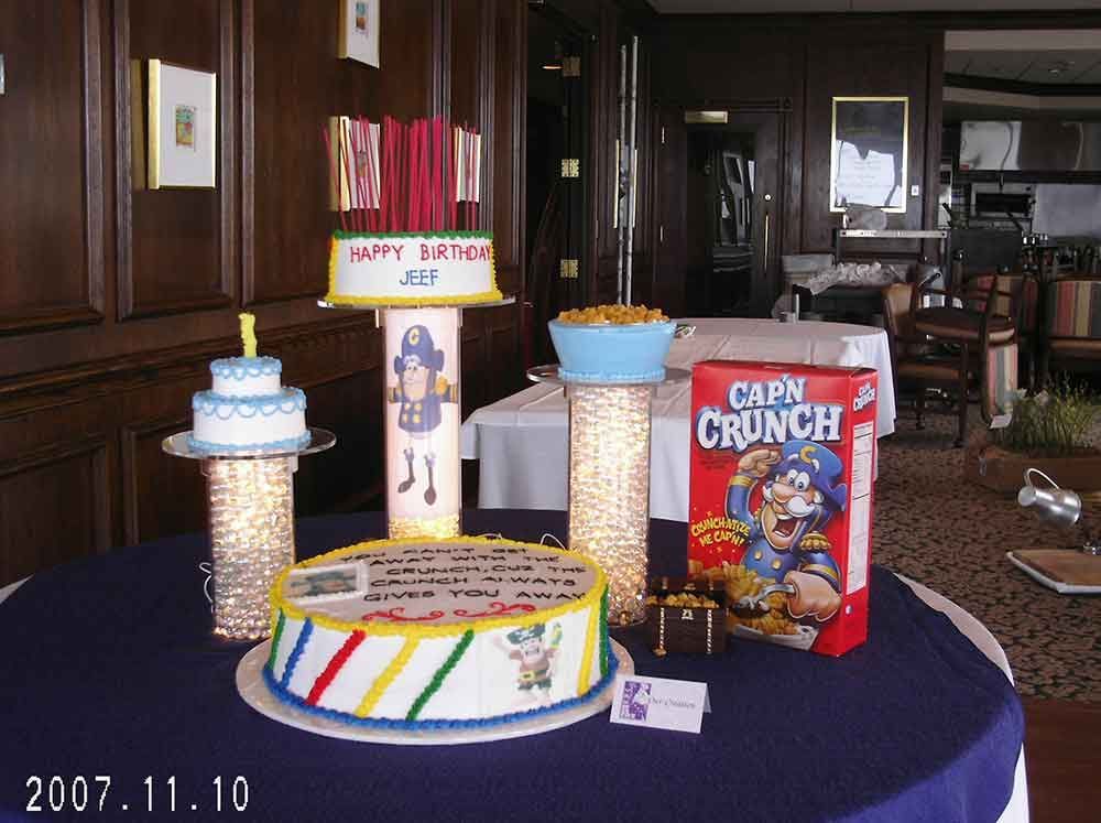 A birthday cake display on a table with several tiers, including a Cap'n Crunch box, set in a wood-paneled room.