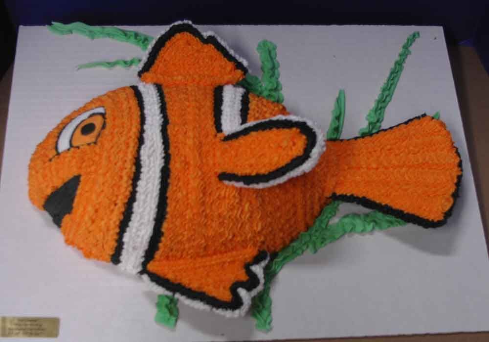 Cake shaped like an orange clownfish with white and black stripes and green seaweed on a white surface.