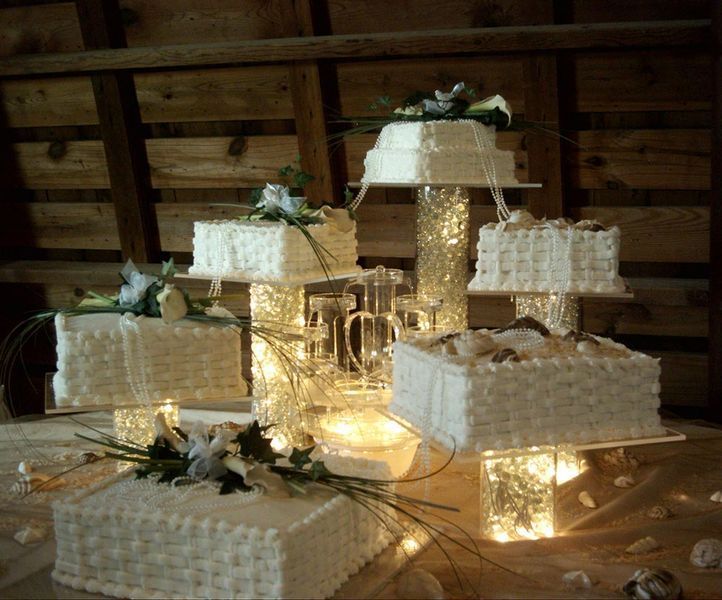 Six-tiered wedding cake display, rectangular cakes with white frosting, decorated with flowers, lit from below.