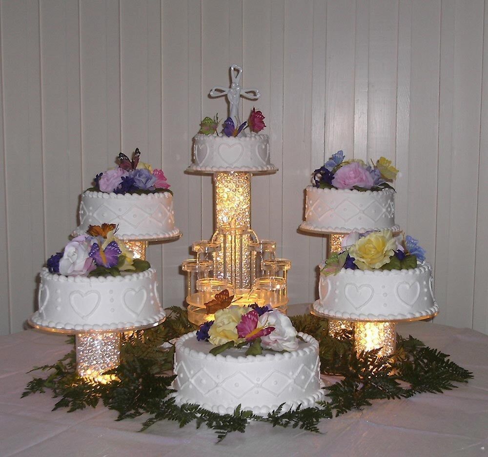 Five-tiered wedding cake display with white cakes, flowers, and a central cross, lit from below.