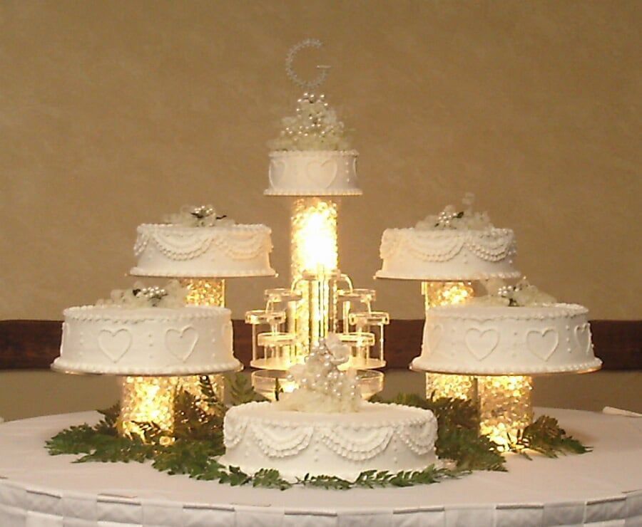 Wedding cake display: multiple tiers with white frosting, lights, and greenery on a round table.