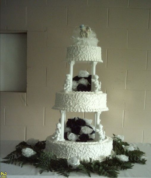 Three-tiered white wedding cake with white pillars and dark and white flower accents, with greenery and white flowers at the base.