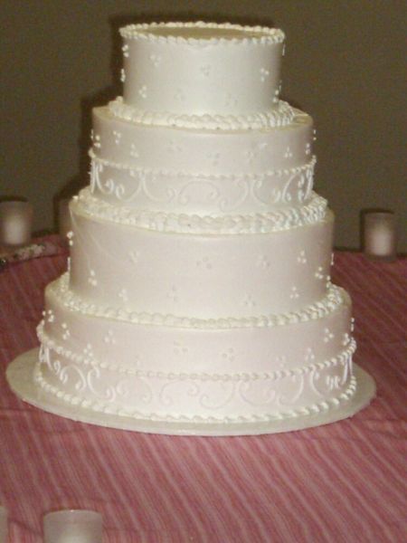 Four-tiered white wedding cake decorated with piped frosting and sitting on a pink tablecloth.