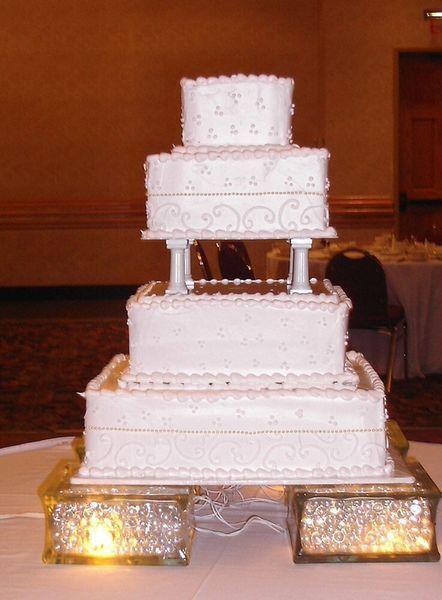 Four-tiered white wedding cake with ornate details, atop lighted decorative blocks.