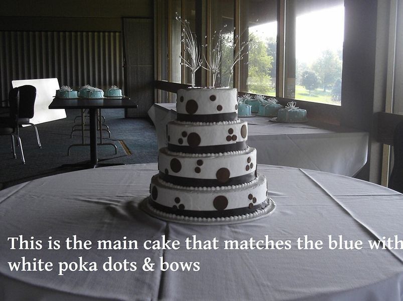 Four-tiered cake with white polka dots and brown accents on a table. The cake matches blue decor.