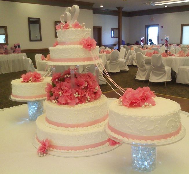 Pink and white tiered wedding cake with floral accents, on a table in a banquet hall.