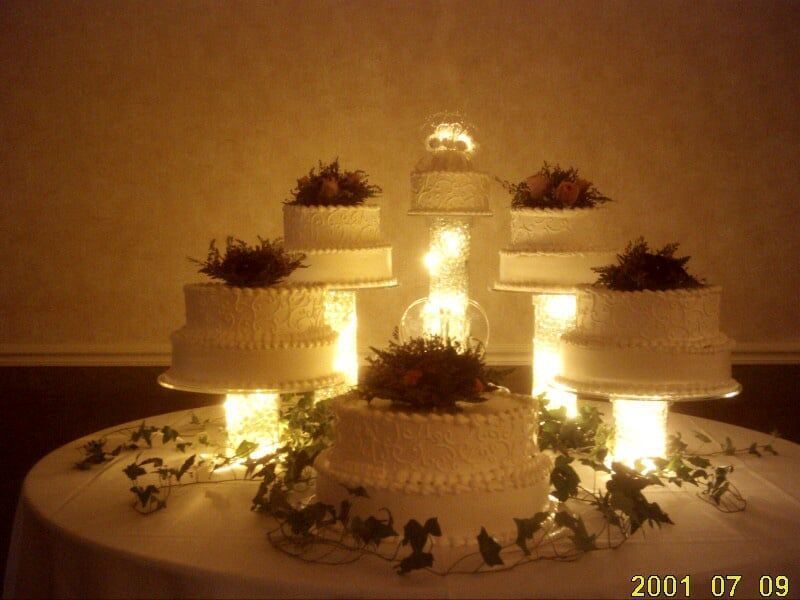 Wedding cake display with multiple tiers, lit from within, arranged on a round table, decorated with flowers and greenery.