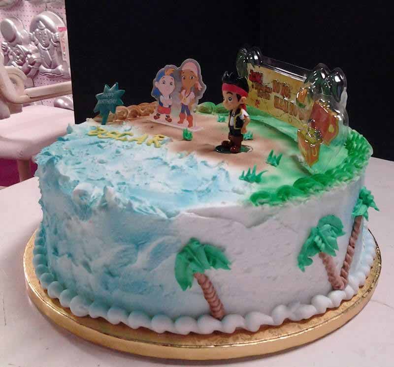Pirate-themed birthday cake with characters and beach scene, light blue frosting, and decorative palm trees.