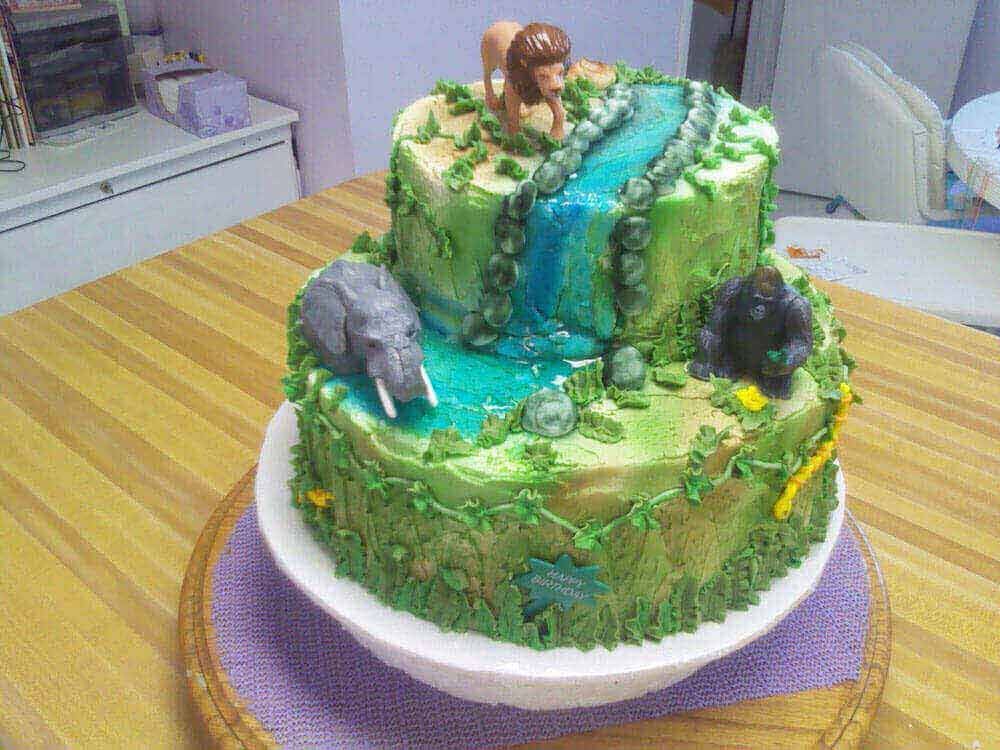 Two-tiered jungle cake with a waterfall, lion, elephant, and gorilla. Green, brown, and blue colors.