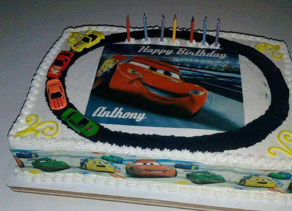 Cars-themed birthday cake with Lightning McQueen image, race track, toy cars, candles, and 