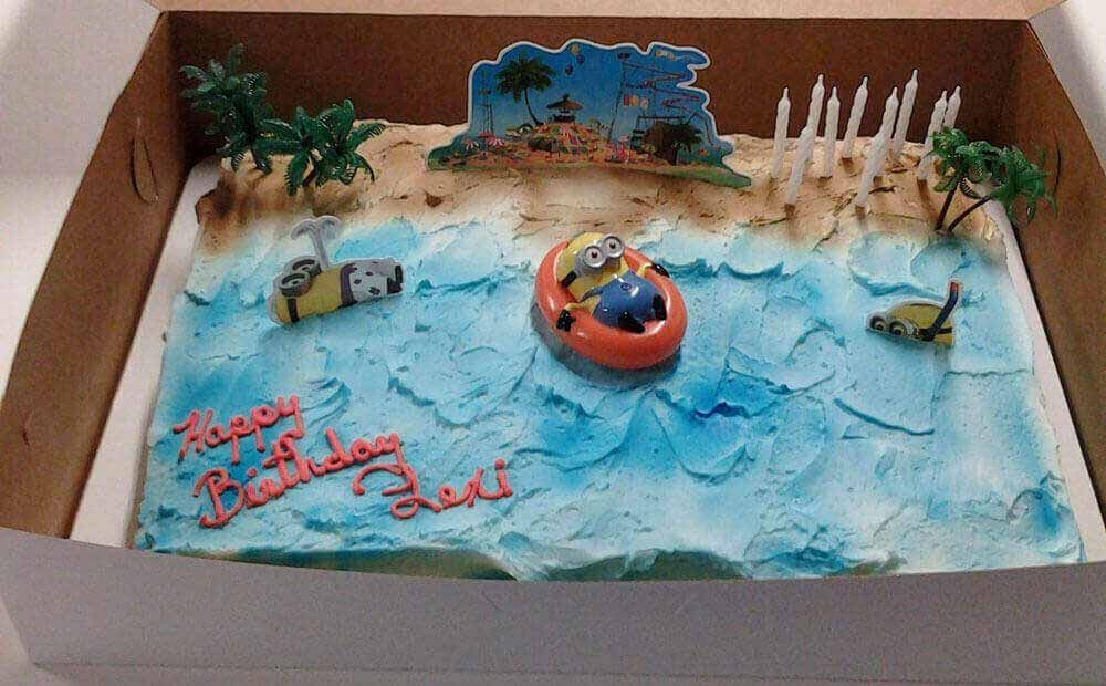 Birthday cake with Minions in a boat on blue frosting “ocean”. Beach scene backdrop. 