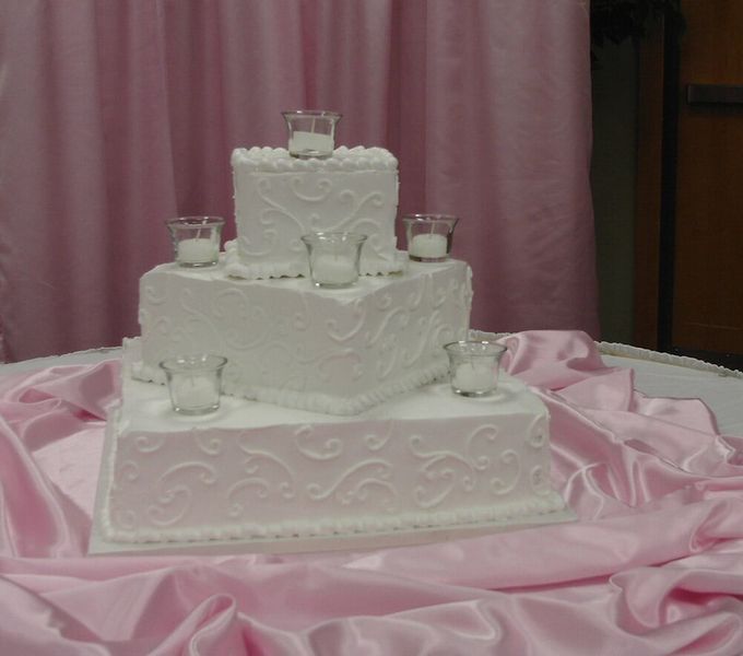 Three-tiered white wedding cake with candleholders, on a pink draped table.