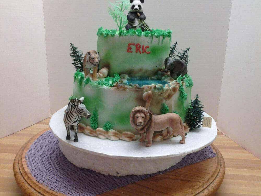 Two-tiered jungle-themed cake with toy animals, including a zebra, lion, panda, and elephant.