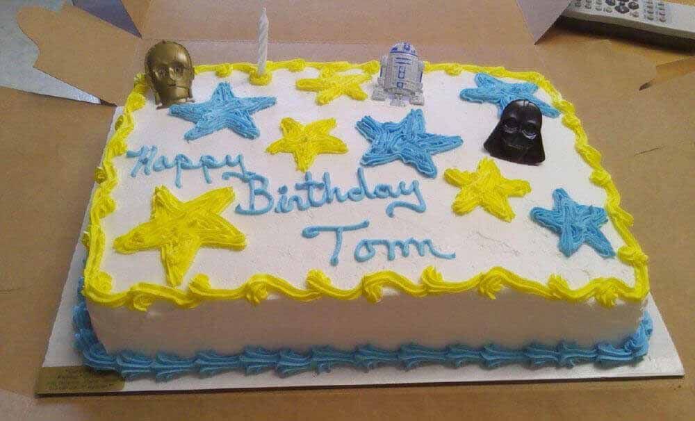 Star Wars themed birthday cake with blue and yellow frosting, figurines, and a lit candle.