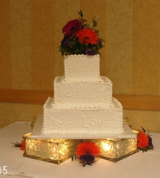 Three-tiered white wedding cake with floral topper, on a lighted crystal-like stand, with the words 