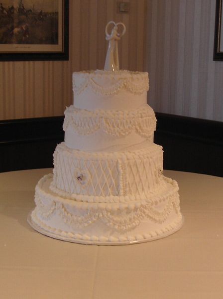 Three-tiered white wedding cake with intricate frosting details and a couple figurine on top, on a white tablecloth.