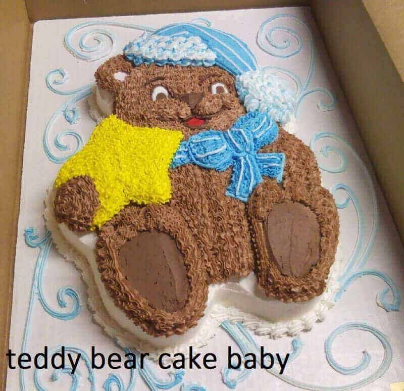 Teddy bear-shaped cake, brown frosting, blue hat and scarf, yellow blanket. White icing border, baby theme.
