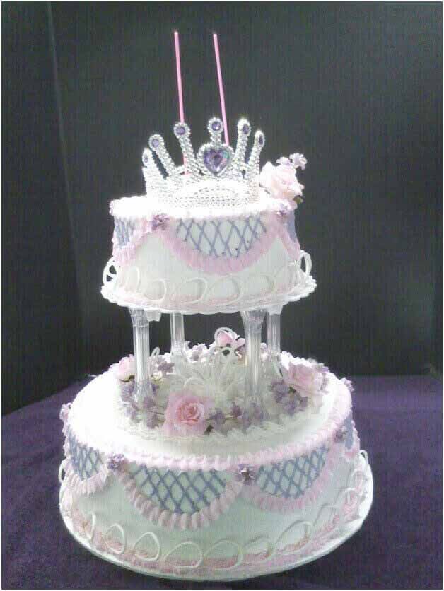 Two-tiered birthday cake, white frosting with pink and blue detailing, topped with a tiara and candles.