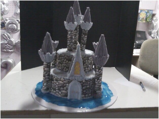 Castle-shaped cake on a plate, blue accents, grey stone-like tower design with purple tops, sitting on a white surface.