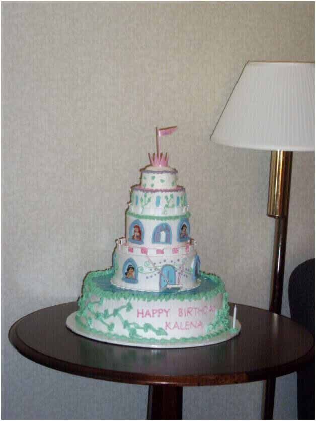 Birthday cake shaped like a castle, on a table near a lamp; green, pink, and blue icing.