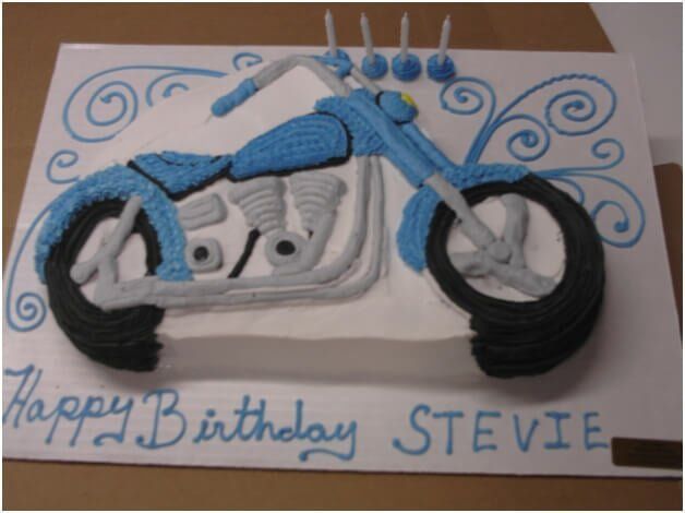 Birthday cake decorated as a blue motorcycle with gray engine, black tires, candles, and blue text that reads “Happy Birthday STEVIE”.