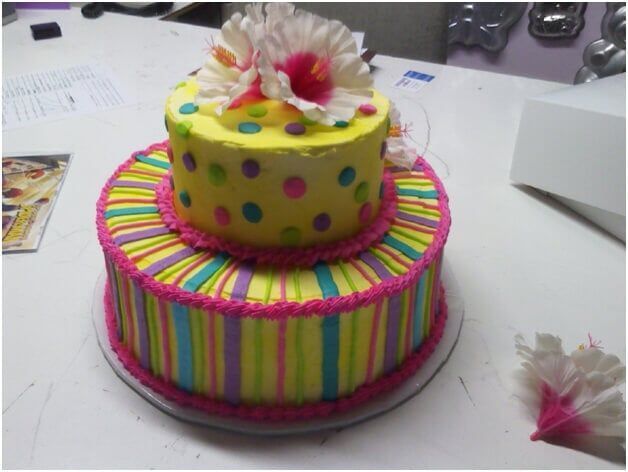 Two-tiered birthday cake with colorful stripes and polka dots, topped with flowers.
