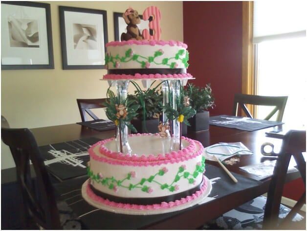 Three-tiered birthday cake with pink and green frosting, a dog on top, on a table.