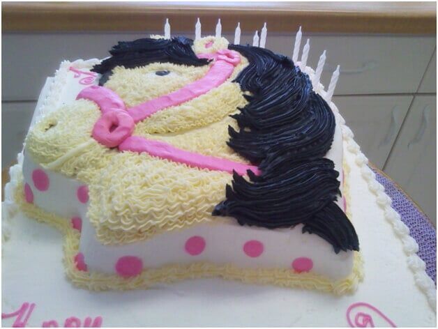 Horse-shaped birthday cake with yellow frosting, black mane, pink bridle, and polka-dot base with lit candles.