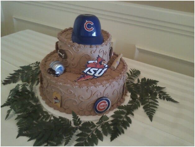Two-tiered cake with blue Cubs baseball cap, ISU logo, and green fern on white tablecloth.