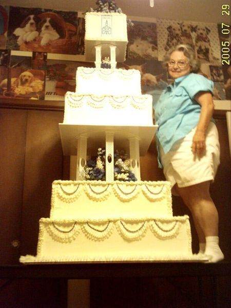 Woman beside a large, multi-tiered white cake with blue accents, displayed indoors near dog photos.