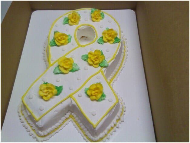 Cake shaped like a yellow ribbon, decorated with yellow roses and white frosting.