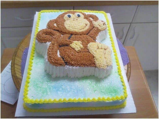 Monkey-shaped cake with brown frosting, a yellow border, and a blue and green background.