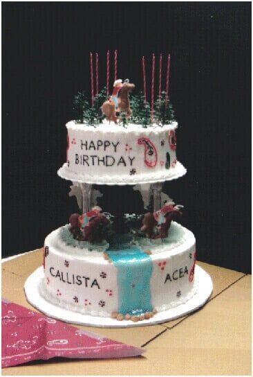 Two-tiered birthday cake with deer, trees, and candles, names 