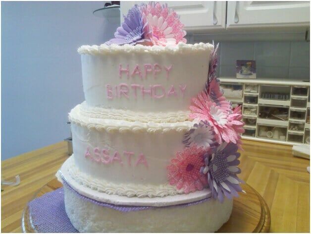Three-tiered white birthday cake with pink and purple flowers. Text reads 
