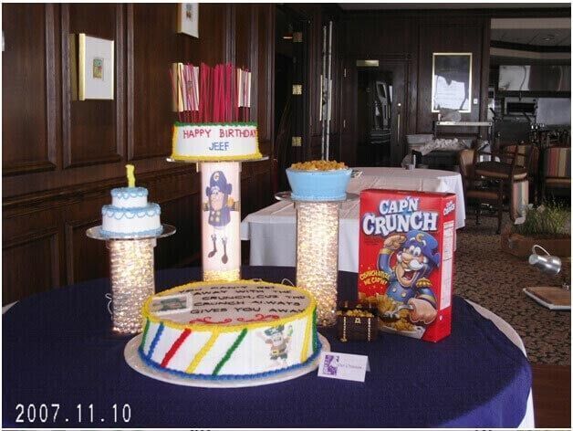 Birthday cake display with multiple tiers, bright colors, cereal box on table with dark blue tablecloth.
