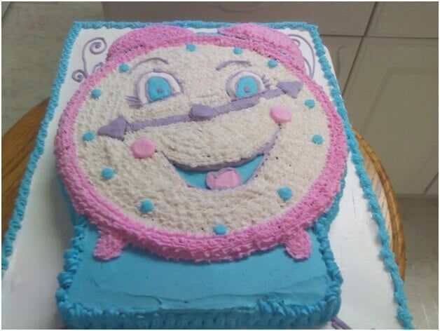 Clock-shaped cake decorated with white, pink, and blue frosting. Clock face has a smiling face.