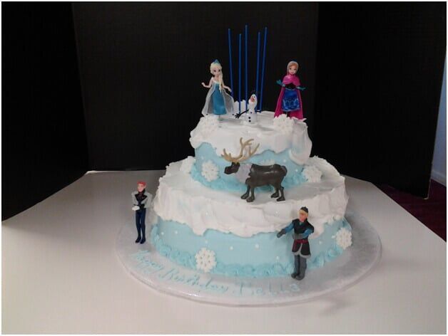 Two-tiered Frozen-themed cake with figurines of Elsa, Anna, Olaf, Kristoff, and a reindeer. Blue frosting and snowflake decorations.