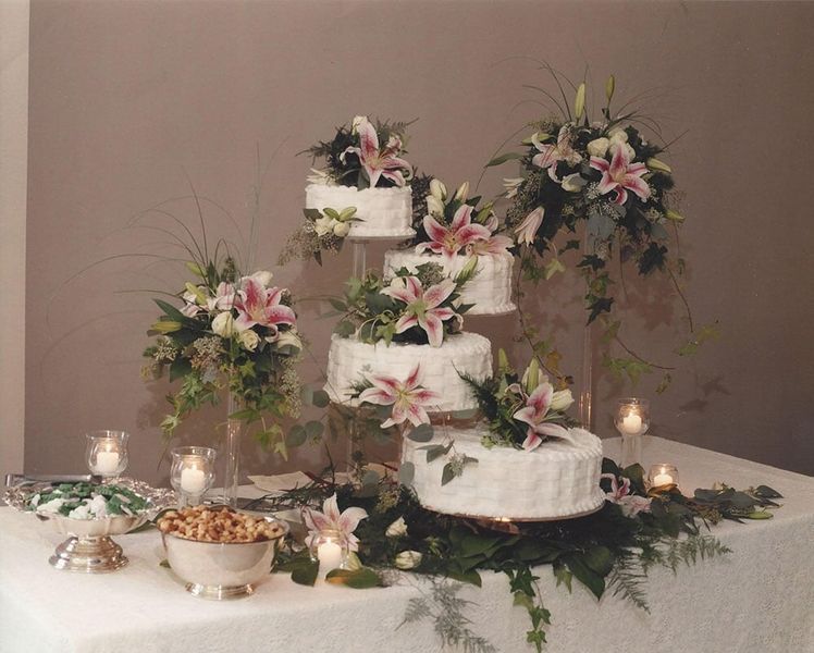 Wedding cake on tiered stand, decorated with lilies and greenery, on a table with candles and snacks.