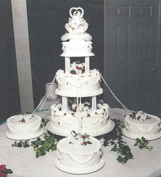 Five-tiered white wedding cake with topper, four smaller cakes around it, decorated with flowers on a table.