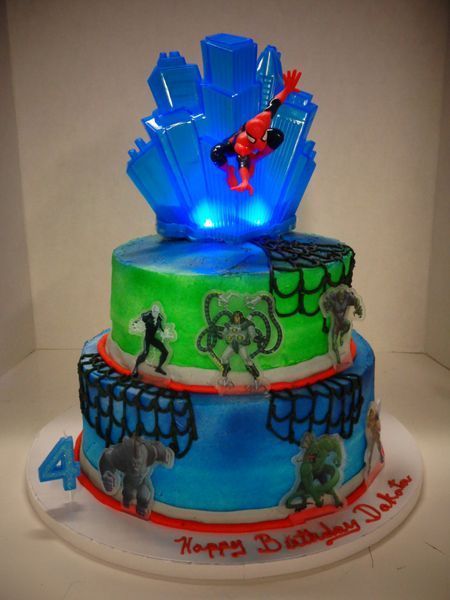 Two-tier Spiderman birthday cake with city skyline topper, figures, blue and green frosting.