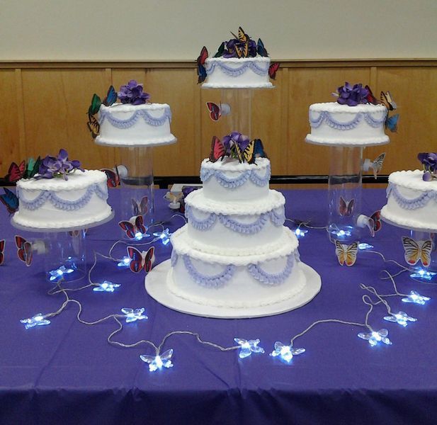 Cake table with tiered white cakes decorated with purple frosting and butterfly accents on a purple tablecloth.