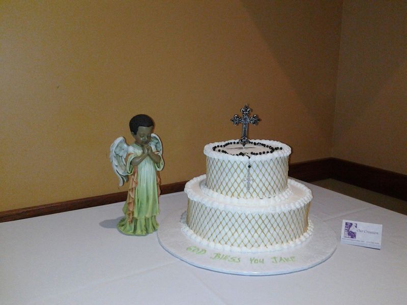 Two-tier white cake with a cross topper and angel statue on a table.