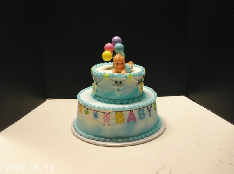 Two-tiered baby shower cake with blue frosting, a baby figurine, and balloons on top, with 