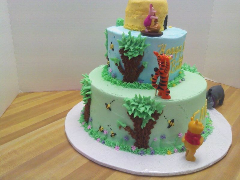 Three-tiered Winnie the Pooh cake with characters and honey pot. Green, blue, and yellow frosting, with trees, grass, and bees.